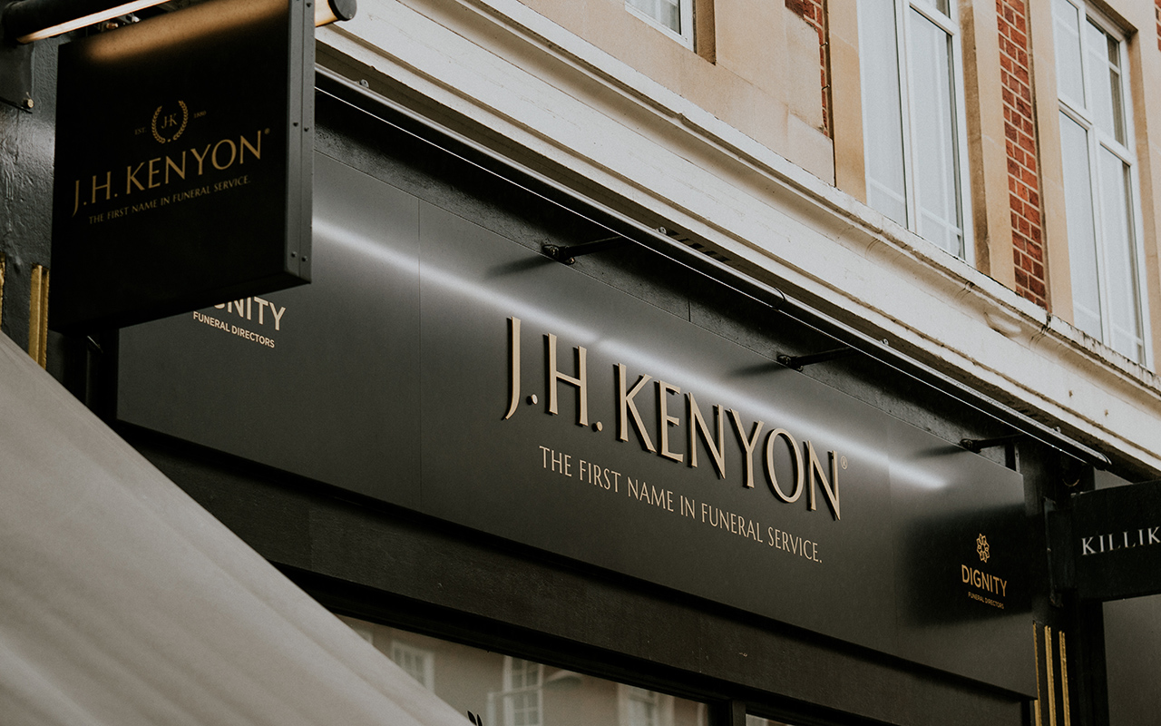 A dark shopfront fascia with gold lettering reading J.H. Kenyon, The First Name In Funeral Service.