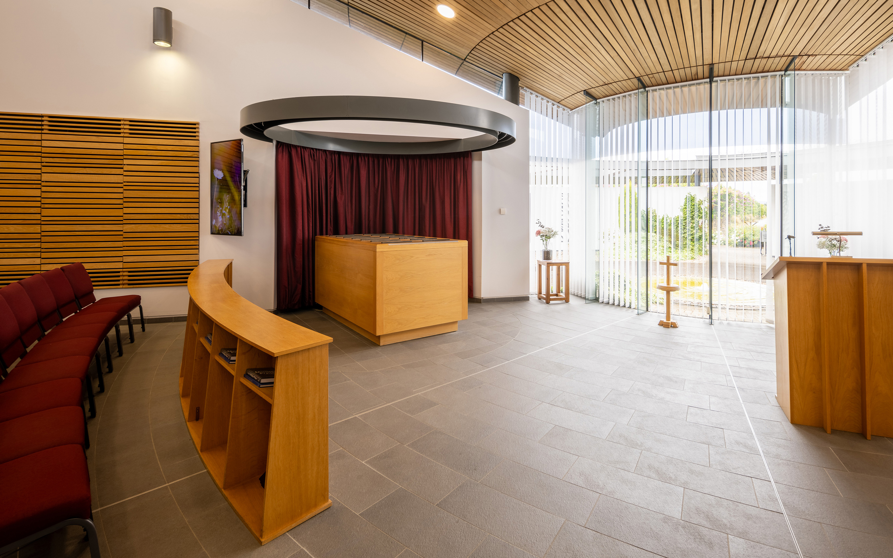 A modern crematorium foyer with a timber ceiling, wooden counters and a glass wall onto countryside.
