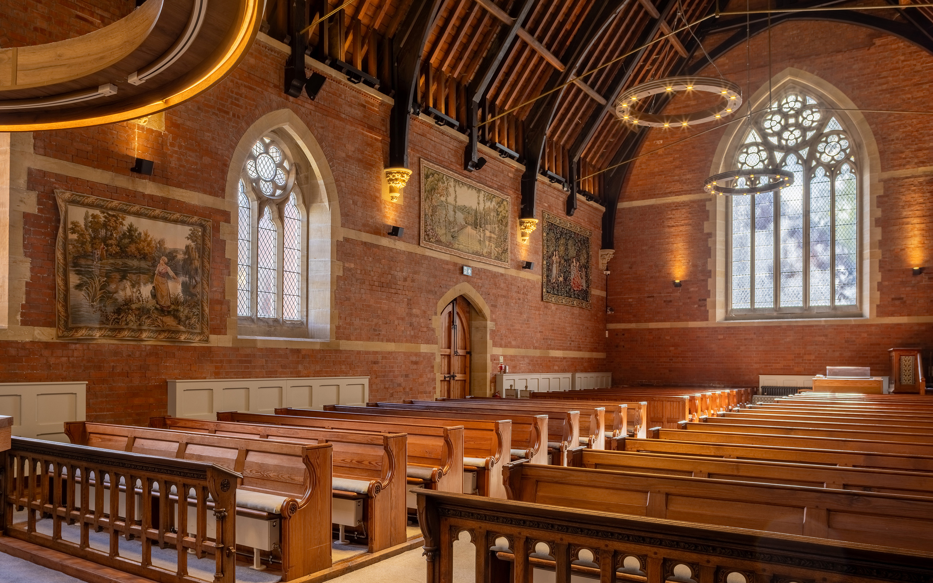 Interior of an empty brick chapel with wooden pews, arched windows and an exposed timber roof.