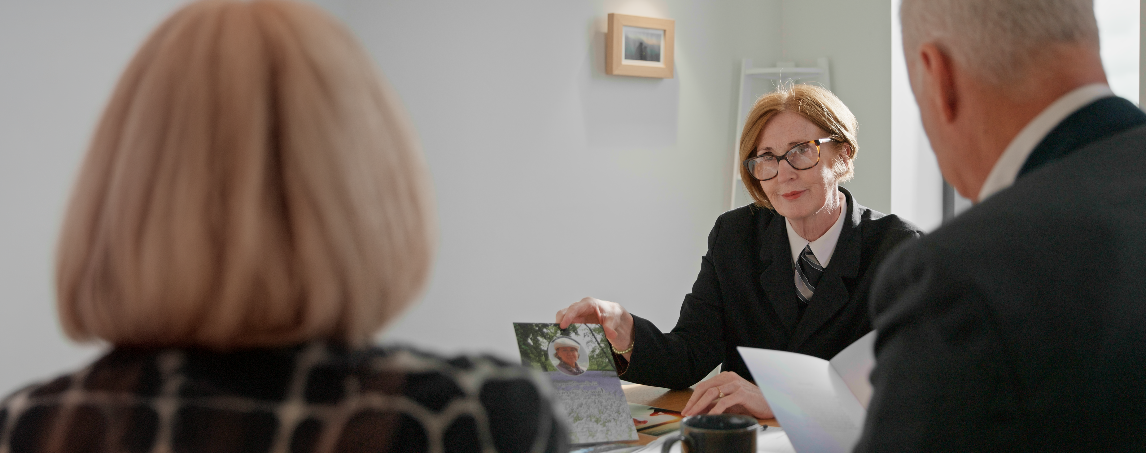 A funeral director at a desk holding a photograph and paperwork, talking to a seated couple.