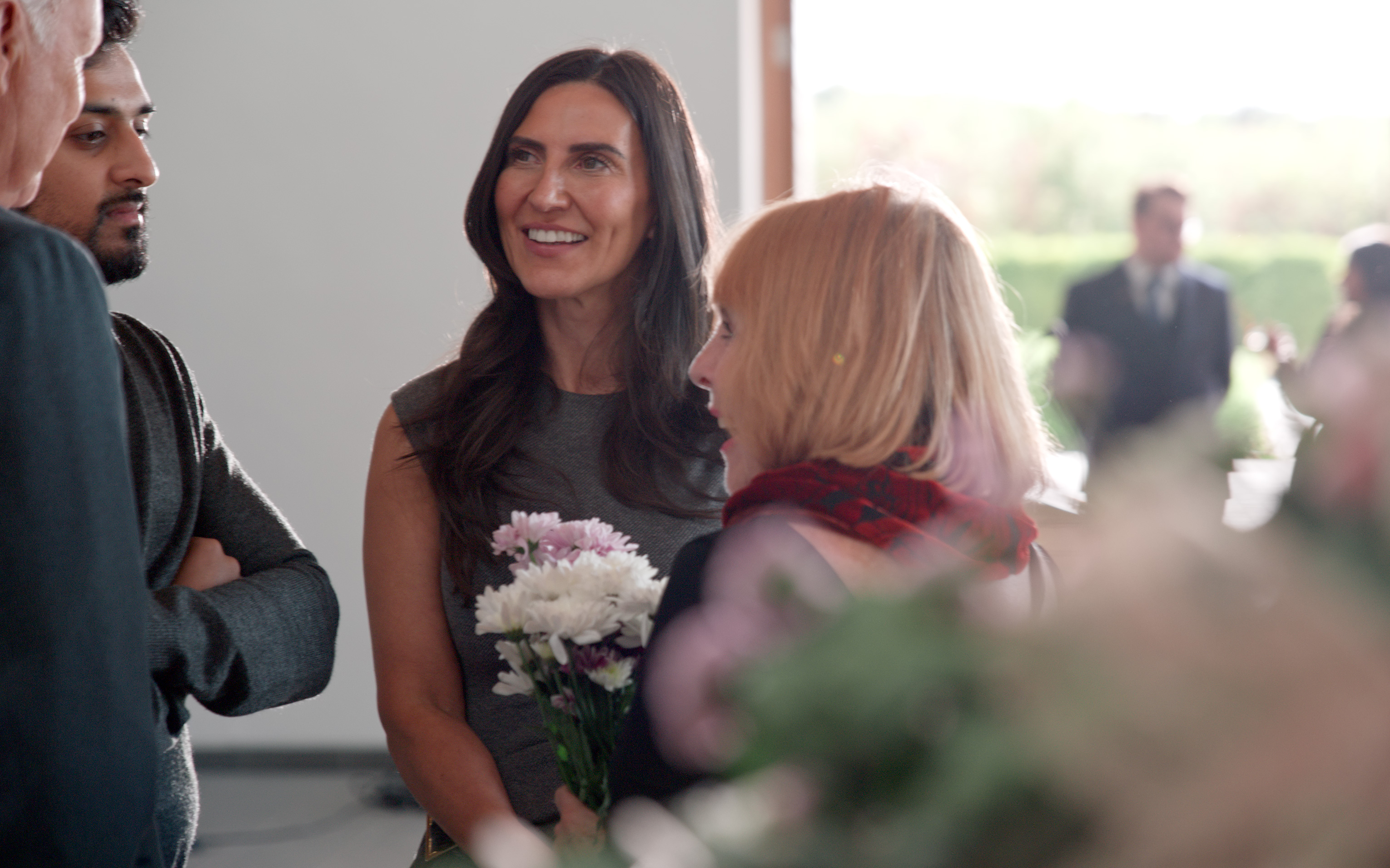 A woman holding white and pink carnations smiles while talking with others at an indoor gathering.