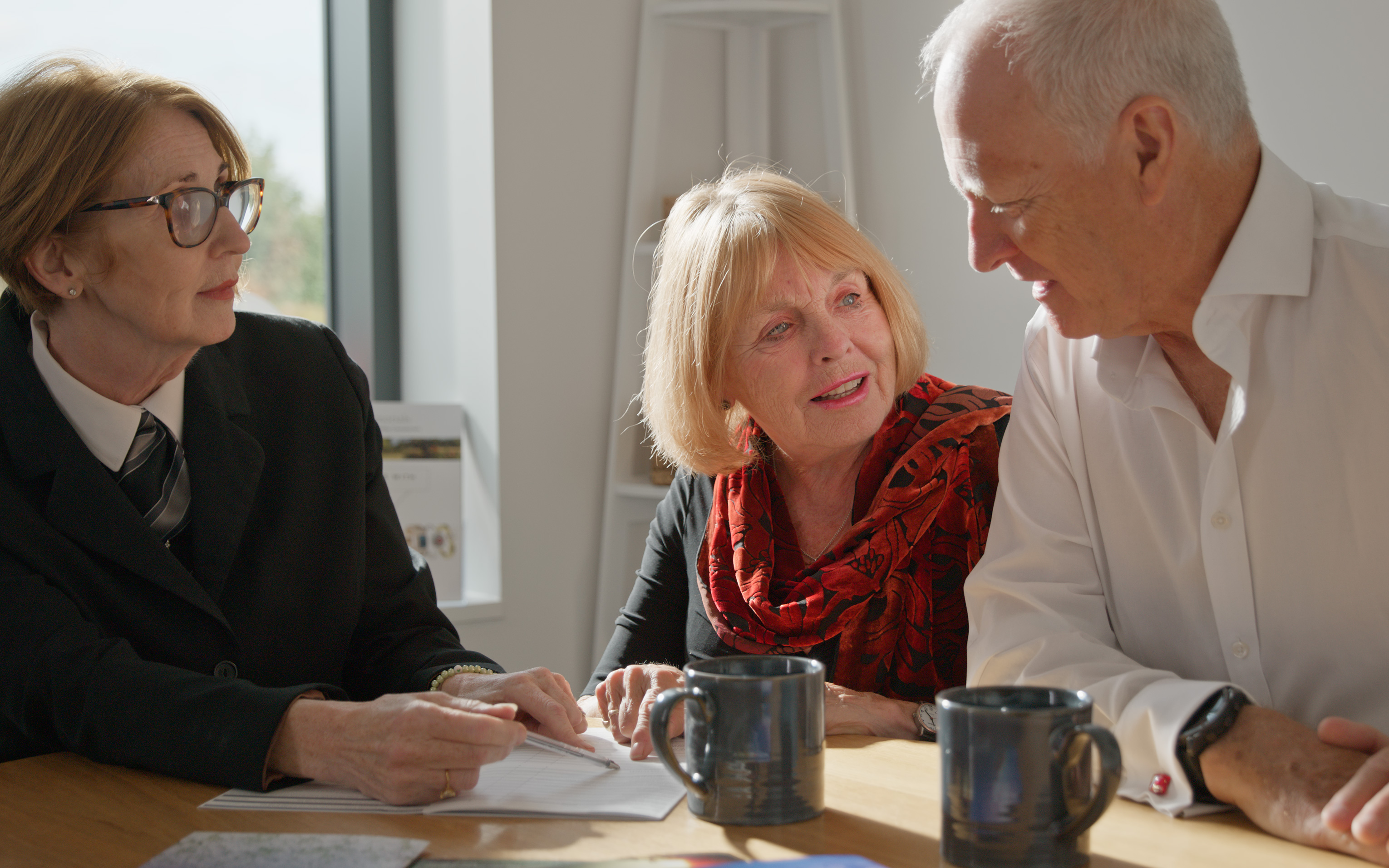 A funeral arranger in a dark suit takes notes with an older couple at a table.