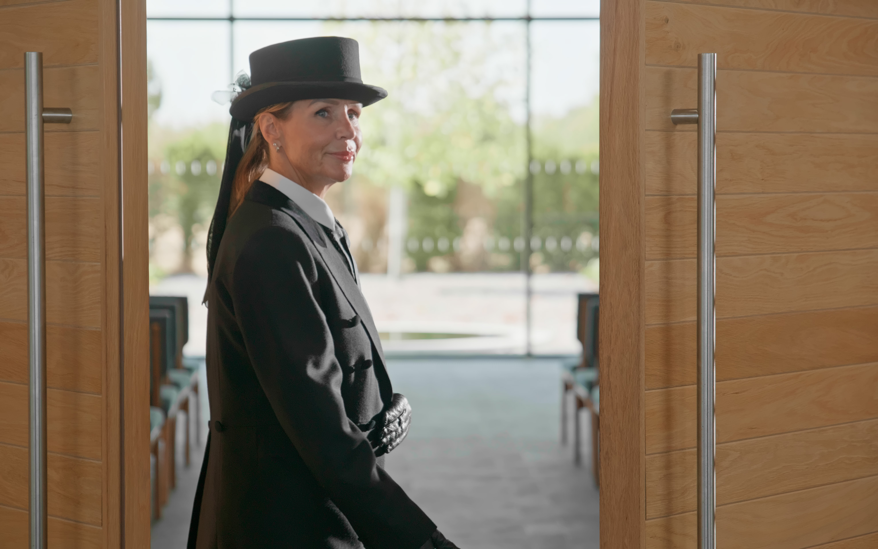 A funeral director in a top hat and long dark coat standing in an open doorway.