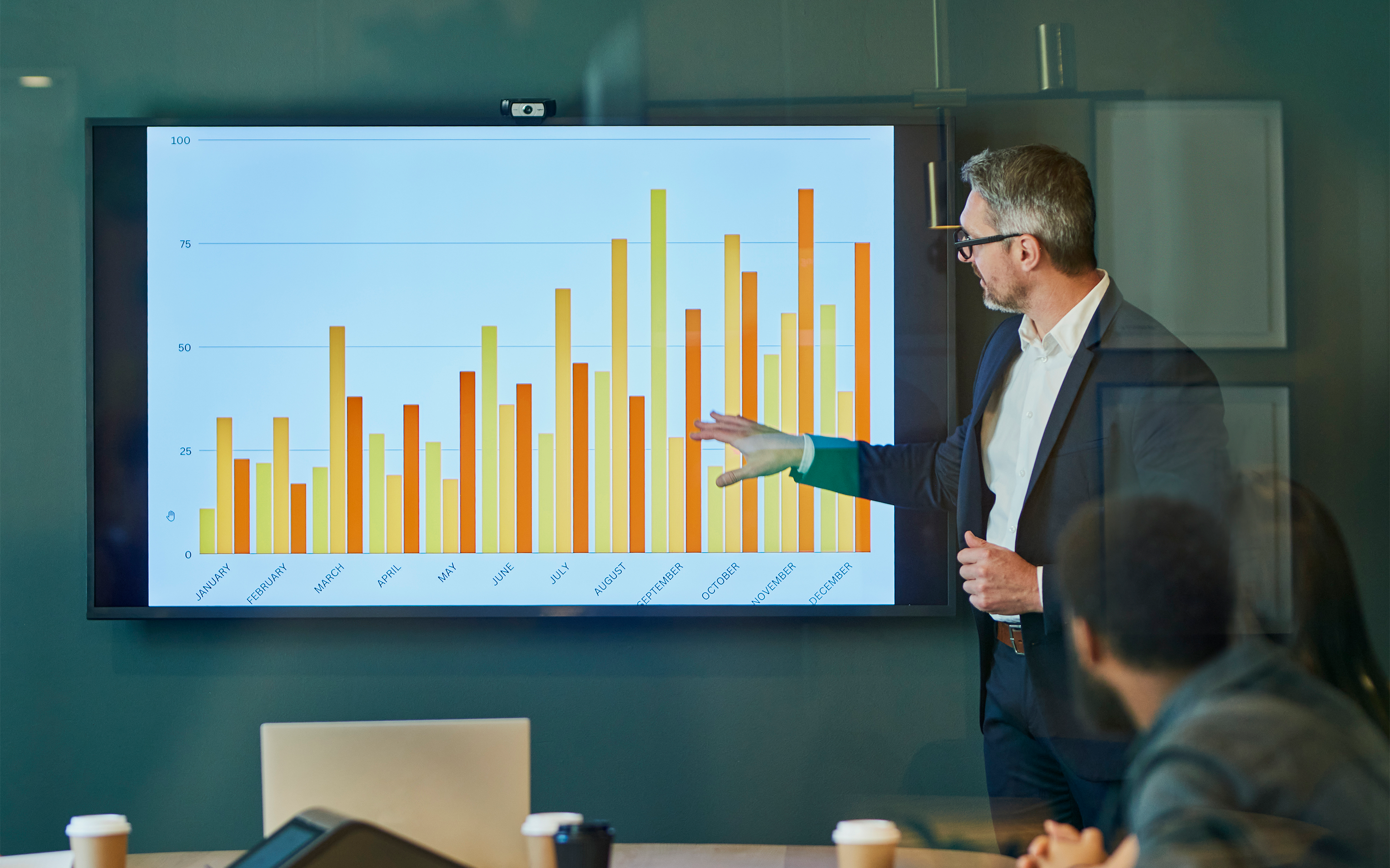 A man presenting a bar chart on a large screen to colleagues in a meeting room.