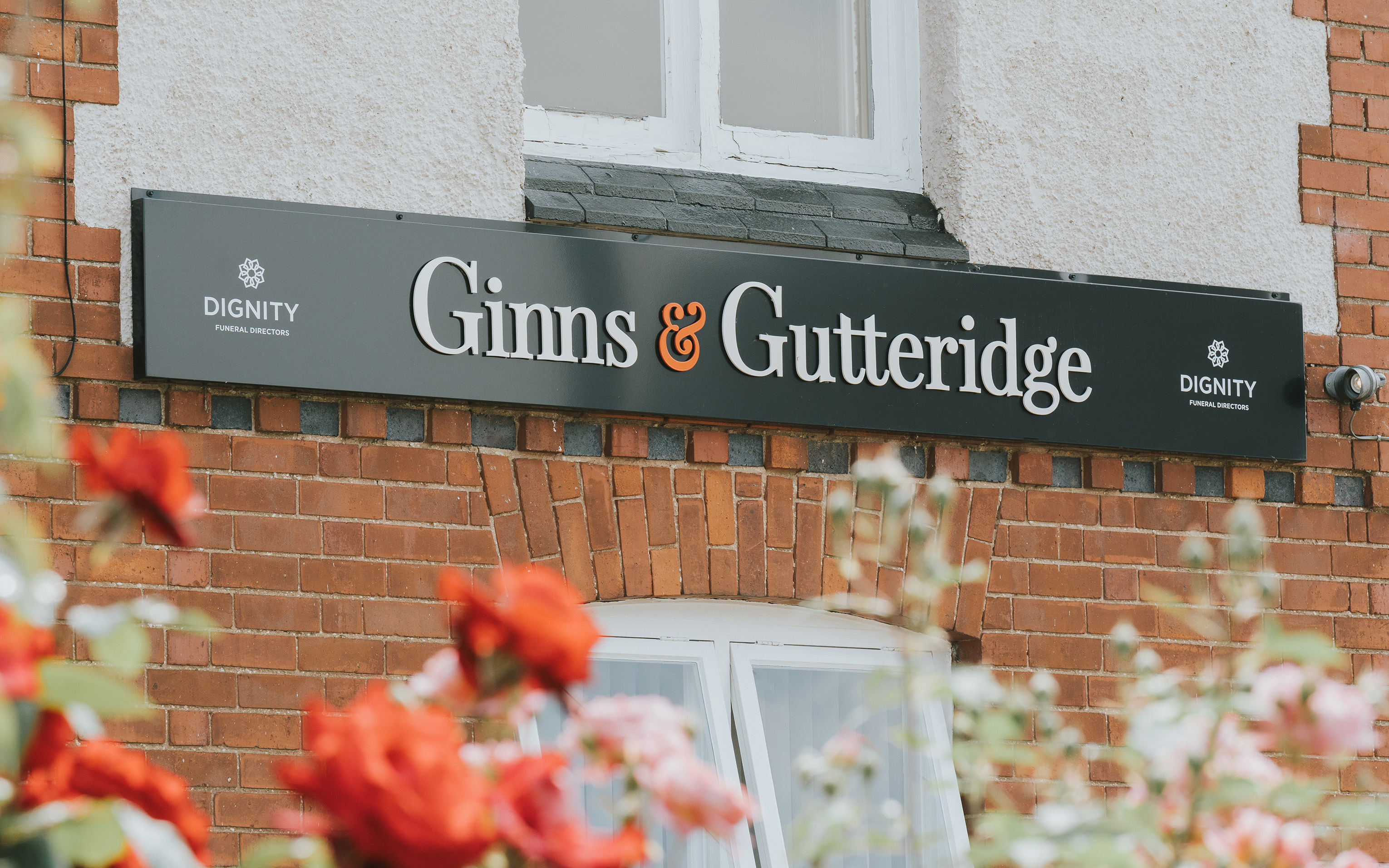 A dark Ginns and Gutteridge sign on a red brick building, with red roses in the foreground.