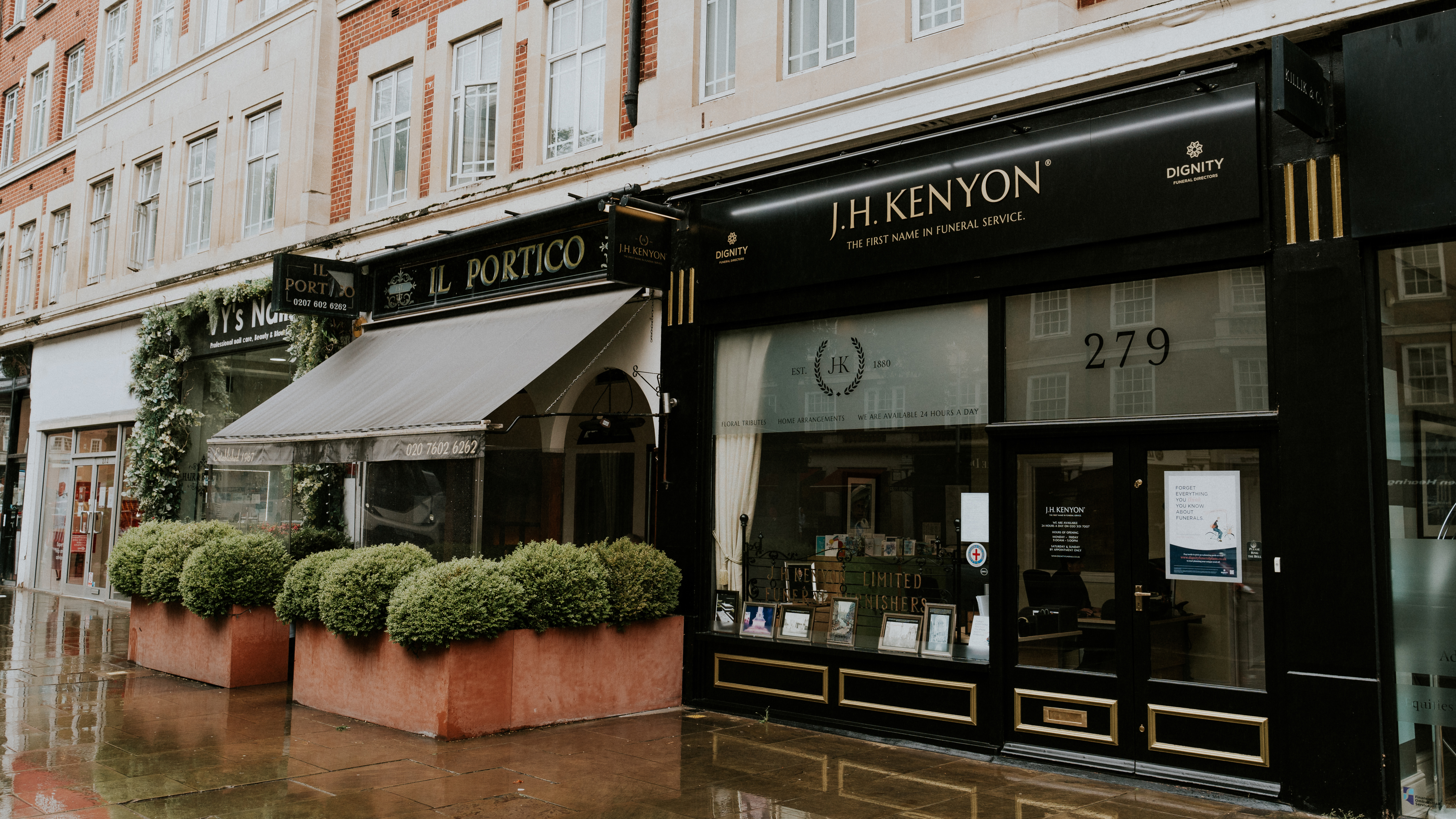 A black shopfront with gold lettering reading J.H. Kenyon Funeral Directors, with clipped hedges outside.
