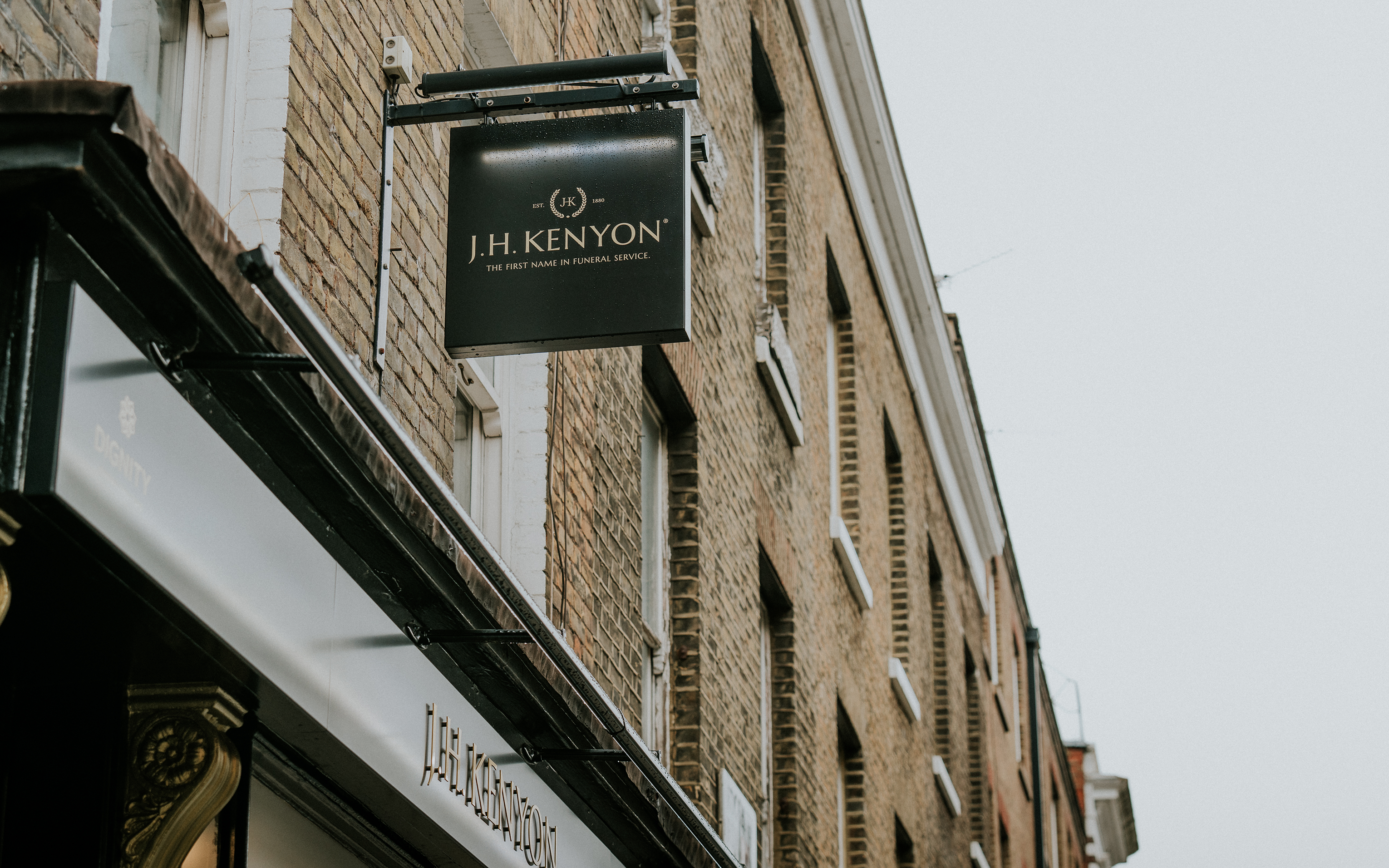 A hanging J.H. Kenyon sign projecting from a brick terraced building against a pale grey sky.