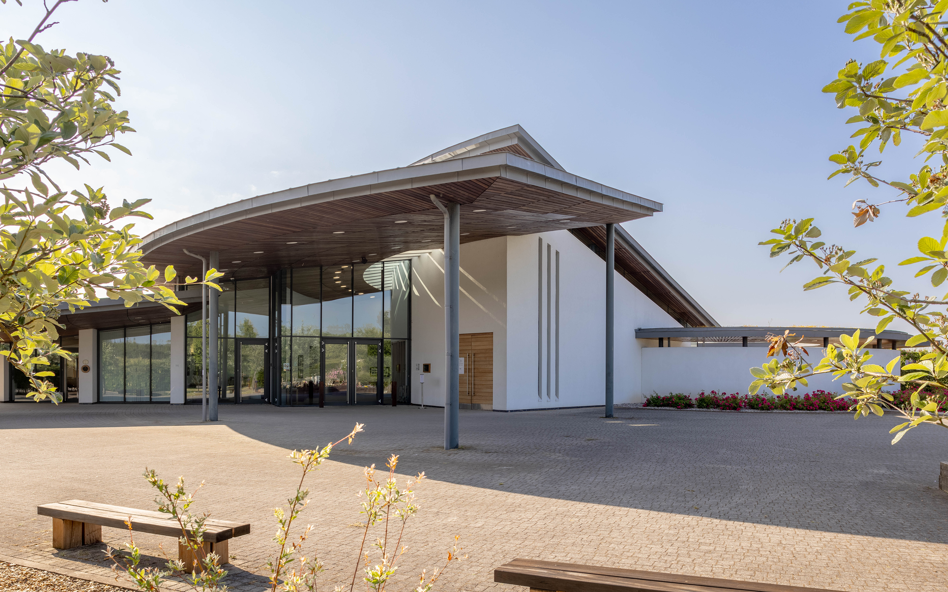 Entrance to a modern building with a wide curved canopy, glass doors and wooden benches.