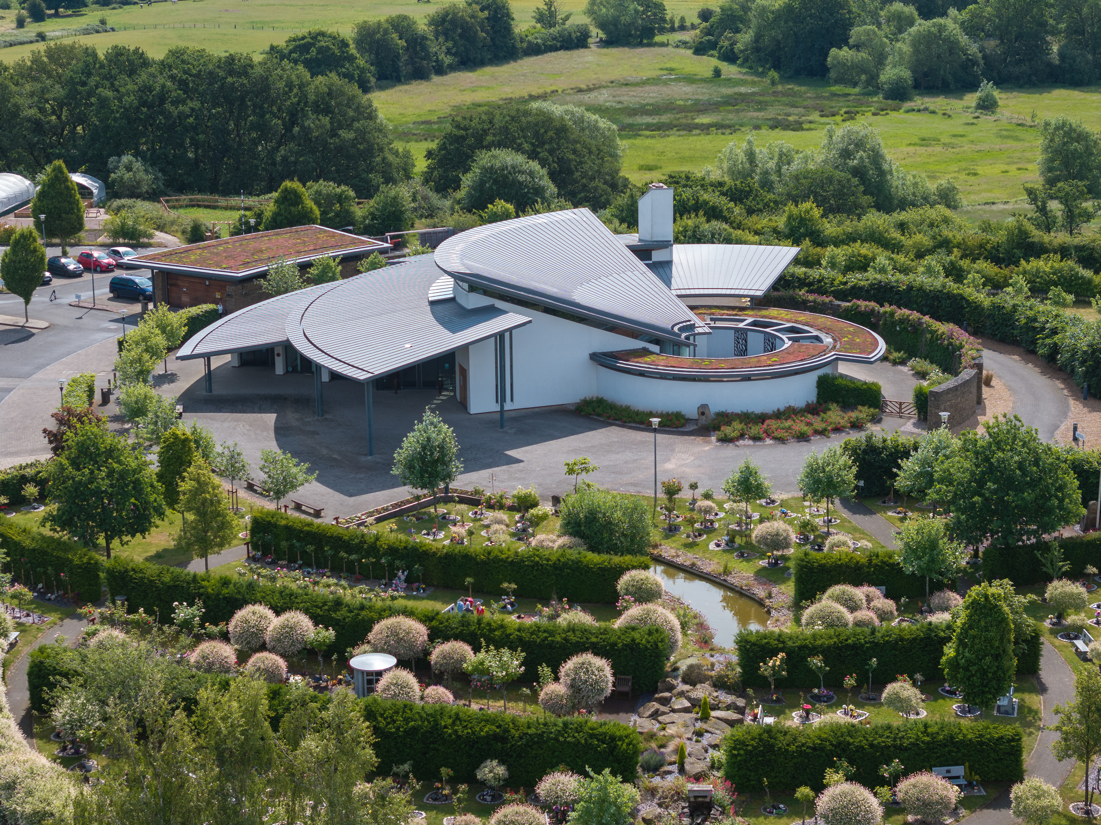 Aerial view of a modern crematorium with a curved roof, set in landscaped gardens and fields.