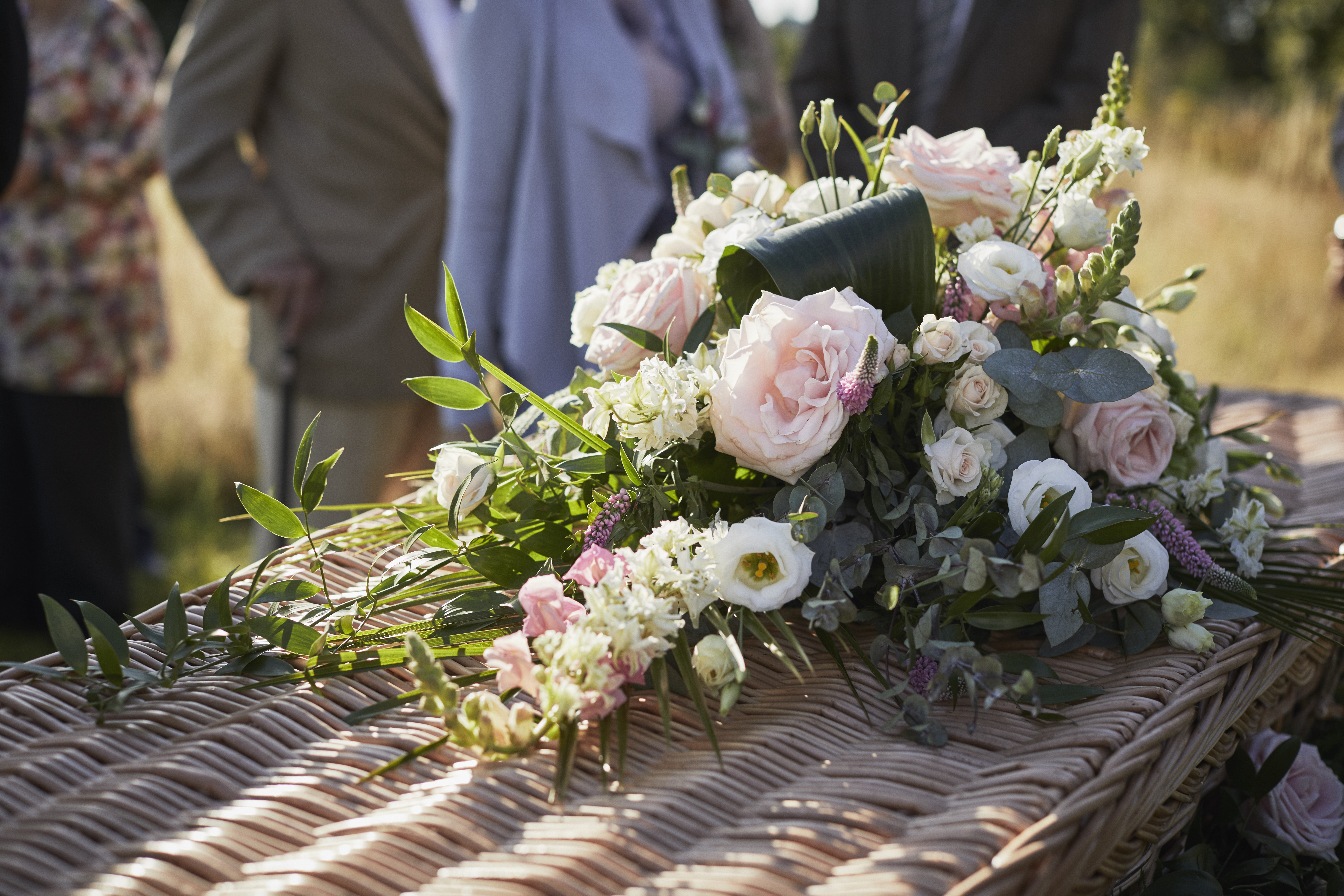A wicker coffin topped with a spray of white and pink roses and eucalyptus, with mourners standing behind.