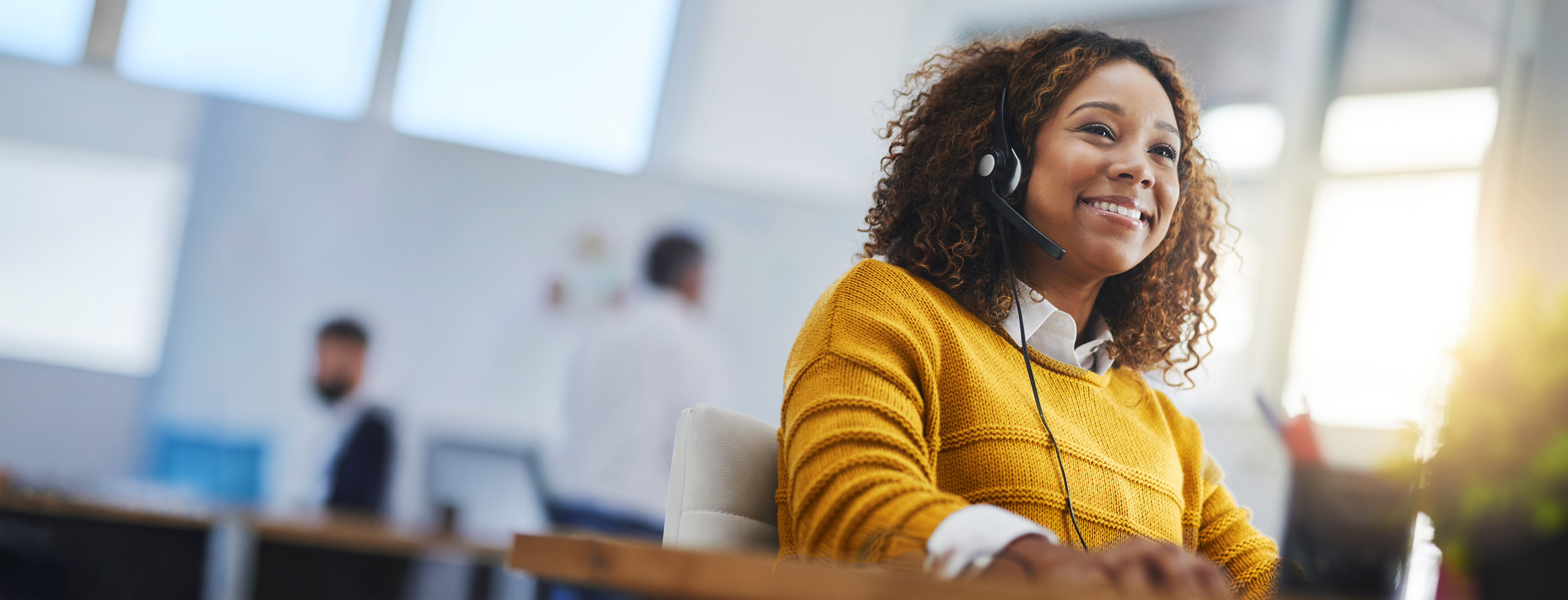 A woman in a yellow cardigan wearing a telephone headset, smiling in an open office.