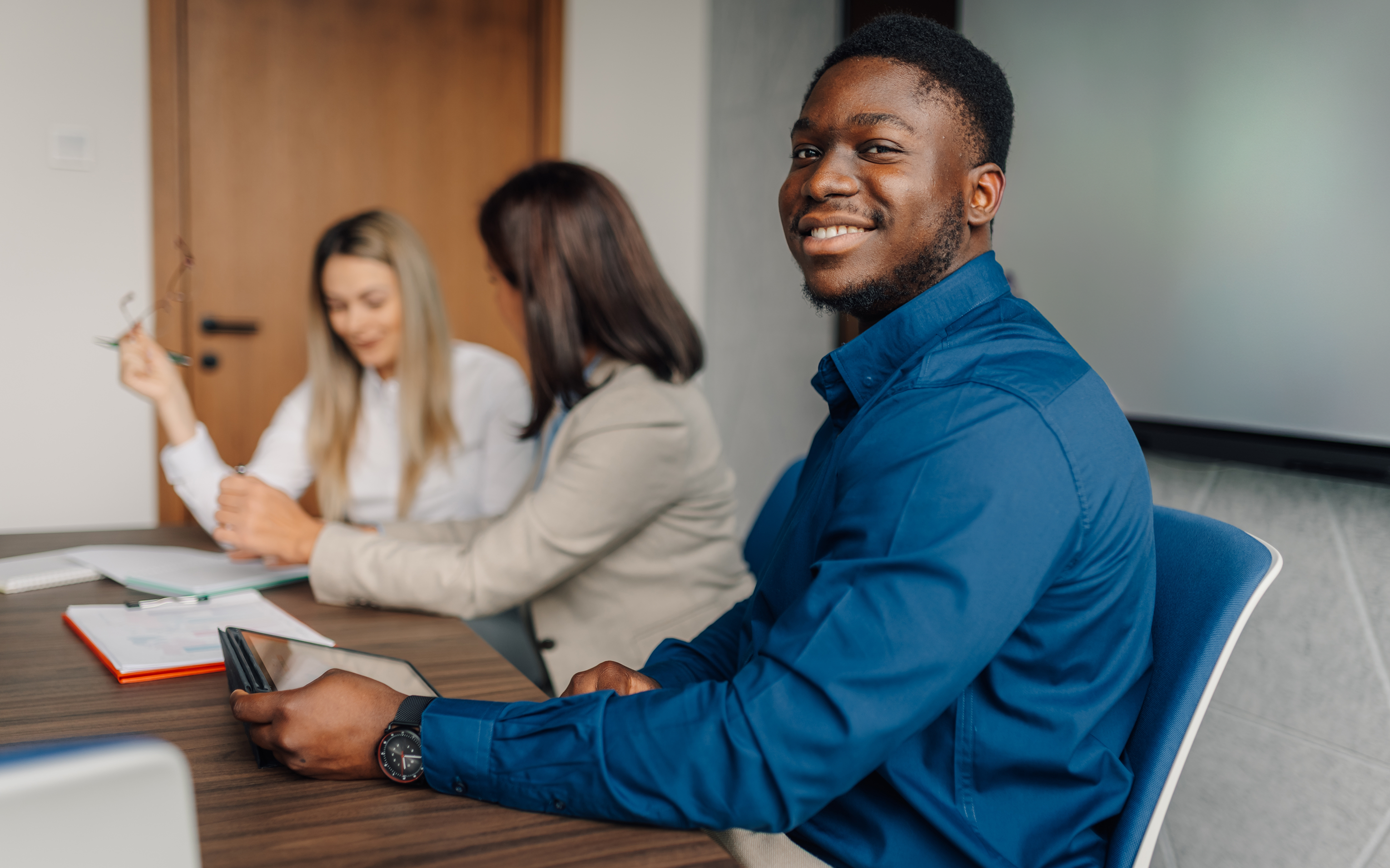 A man in a blue shirt smiles at a meeting table while two women talk behind him.